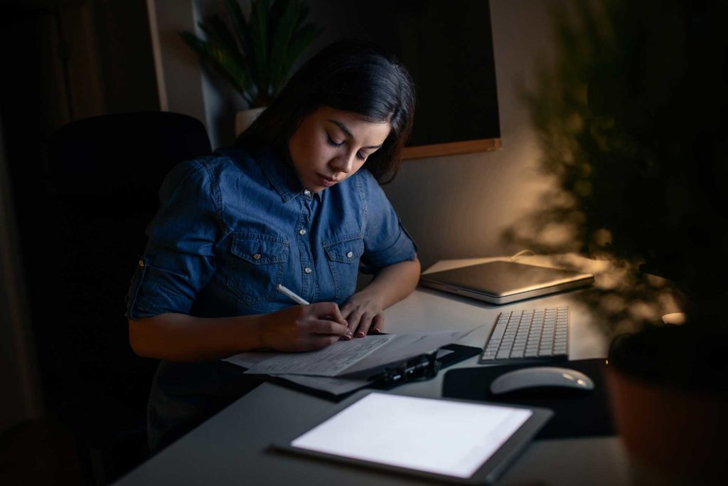 Young woman sits writing at her desk in dim lite room